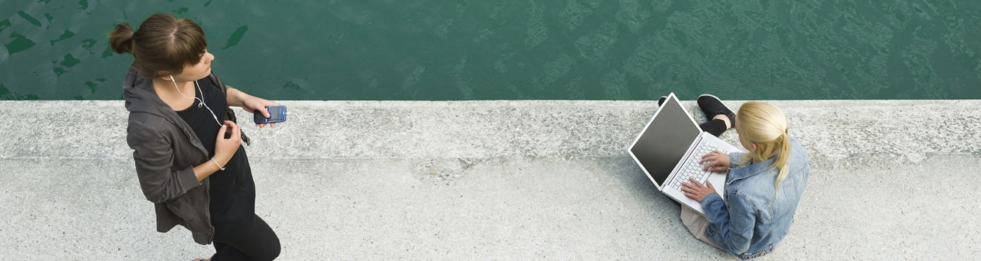 Woman sitting by water on laptop with another woman walking by