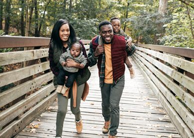 Parents holding small children walking across wooden bridge in forest.